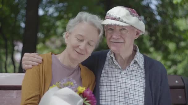 Retrato de una pareja madura enamorada sentada en un banco en el parque. La mujer adulta sostiene un hermoso ramo de flores mientras su anciano esposo la abraza. Tierna relación pareja adulta al aire libre . — Vídeos de Stock