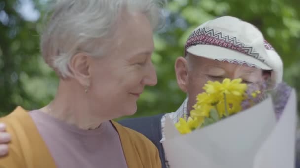 Parkta bir bankta oturan aşık olgun bir çiftin portresi. Yetişkin kadın ve yaşlı kocası bir buket çiçek kokluyorlar. İhale ilişki yetişkin çift açık havada. — Stok video