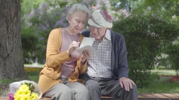 Портрет очаровательная взрослая пара, смотрящая старые фотографии, вспоминающие счастливые моменты, сидя на скамейке в парке. Зрелая влюбленная пара отдыхает в солнечный теплый весенний день на открытом воздухе . — стоковое видео