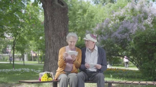 Retrato linda pareja adulta mirando fotos viejas recordando momentos felices sentados en un banco en el parque. Pareja madura enamorada descansando en un soleado y cálido día de primavera al aire libre . — Vídeo de stock