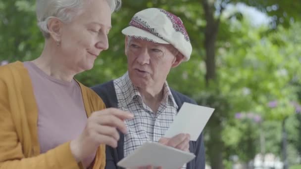 Close-up portret schattig volwassen paar op zoek naar oude Foto's het herinneren van gelukkige momenten zittend op een bankje in het Park. Volwassen paar in liefde rusten op een zonnige warme lente dag buitenshuis. — Stockvideo