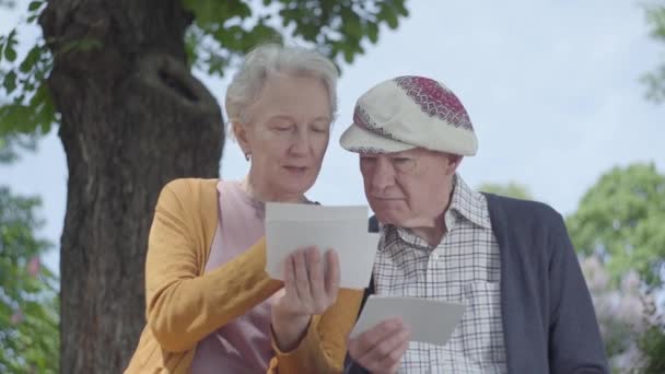 Portrait adorable mature woman and man looking old photos remembering happy moments sitting on a bench in the park. Adult couple in love resting on a sunny warm spring day outdoors. — Stock Video