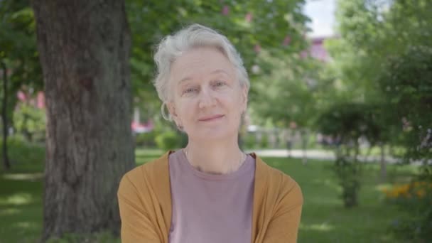 Retrato de una linda anciana con el pelo gris sonriendo en el parque verde increíble. Adorable abuela madura descansando en un soleado y cálido día de primavera al aire libre. Cambiar las emociones en la cara de una mujer — Vídeos de Stock