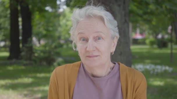 Portret van schattige smilling oude vrouw met grijs haar en blauwe ogen zittend in het groene verbazingwekkende Park. Schattig volwassen oma rusten op een zonnige warme lente dag buitenshuis. — Stockvideo