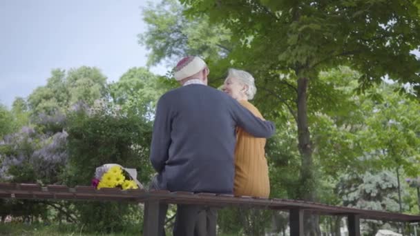 Bak av gammal man sitta i bänken i parken med bukett gula blommor och kram kvinna med grått hår. Kvinna sätta huvudet på män skuldra — Stockvideo