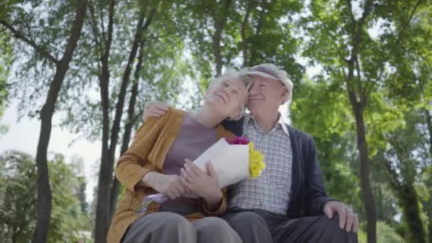 Portret dojrzałej pary w miłości siedzi na ławce w parku. Dorosły kobieta trzyma piękny bukiet kwiatów, podczas gdy jej starszy mąż uściski jej. Delikatny związek urocza para na dworze. — Wideo stockowe