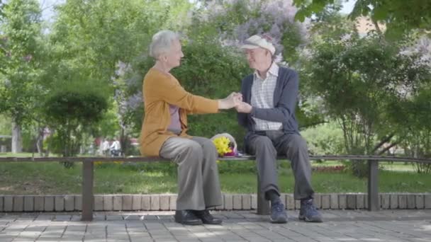 Portrét rozkošné stařenky s kyticí žlutými květy sedí na lavičce se starým mužem a drží se v parku. Nabídka vztahu dospělí manželé venku. — Stock video