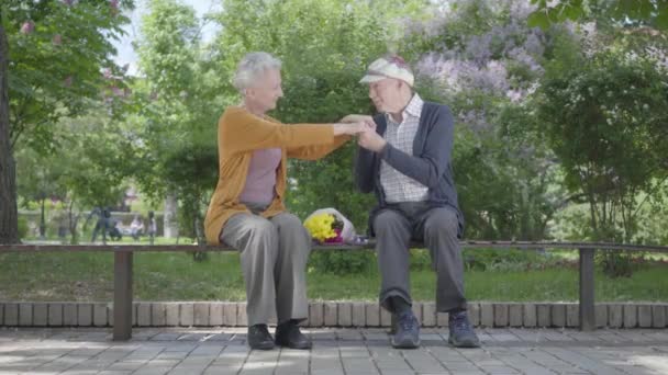 Porträtt gammal kvinna med bukett gula blommor sitta i bänken med en gammal man och hålla händerna i parken. Anbuds förhållande vuxen par utomhus. — Stockvideo