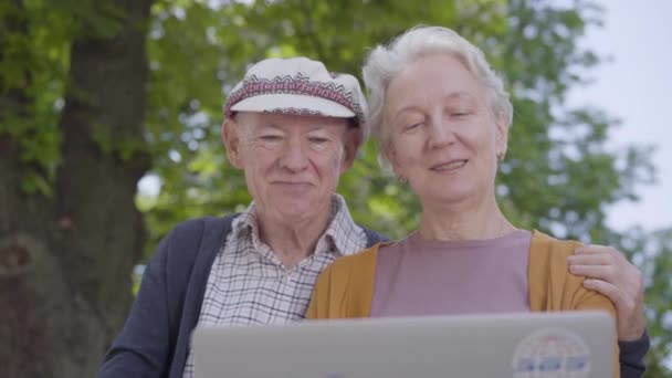 Retrato linda anciana con el pelo gris y el hombre viejo en la tapa sentado en el banco en el hermoso parque y hablando con los niños de la computadora portátil . — Vídeo de stock