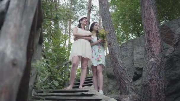 Dos mujeres jóvenes en trajes cortos con flores de pie en las escaleras y mirando amzing bosque, árboles, plantas. Chicas guapas pasean fuera de la ciudad. Lindas novias pasando el fin de semana juntas al aire libre . — Vídeo de stock