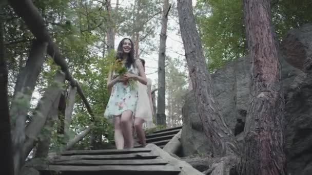 Zwei junge Frauen in kurzen Kleidern mit Blumen gehen die Treppe hinunter und schauen sich Wald, Bäume, Pflanzen an. Verblüffende Sicht. — Stockvideo