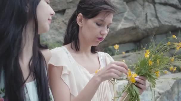 Dos hermosas mujeres con pelo negro corona de flores trenzada cerca de piedra enorme con plantas verdes. Adorables novias pasando el fin de semana juntas caminando al aire libre . — Vídeos de Stock