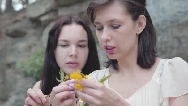 Two beautiful women with black hair braid flower crown near huge stone with green plants. — Stock Video