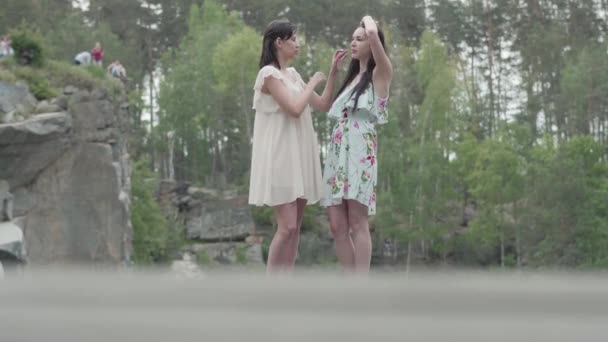 Портрет двох чарівних молодих жінок носить літні сукні, що стоять на вершині скелі і дивляться на дивовижний вид на природу. Милі дівчата виходять за місто. Чарівні подружки проводять вихідні — стокове відео