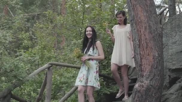 Zwei junge Frauen in kurzen Kleidern mit Blumen gehen die Treppe hinunter und schauen sich Wald, Bäume, Pflanzen an. Verblüffende Aussicht. Zeitlupe. — Stockvideo