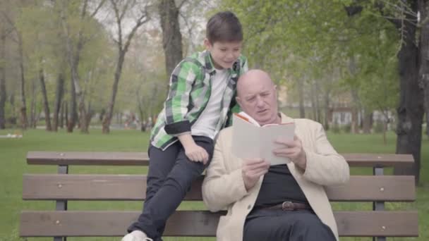 かわいいおじいちゃんと愛らしい孫がベンチの公園に座って、老人が少年のために本を読んで。世代の概念。フレンドリーな家族。屋外レジャー。おじいちゃんと孫 — ストック動画