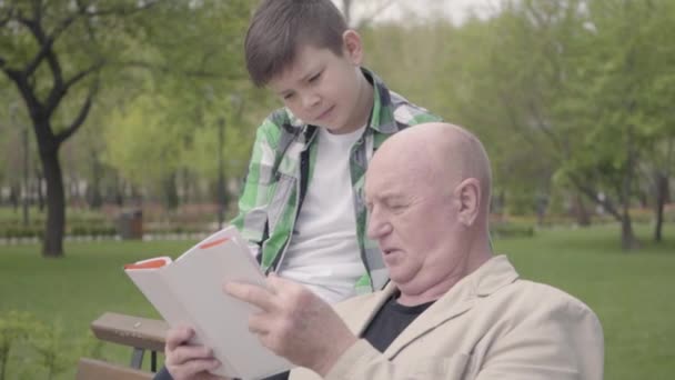 Portret cute dziadek i adorable wnuka siedzi w parku na ławce, staruszek czytając książkę dla chłopca. Koncepcja pokoleń. Rodzinna obsługa. Wypoczynek na świeżym powietrzu. Dziadek i wnuk — Wideo stockowe