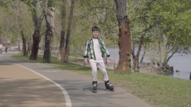 Aranyos fiú fényes ruhákat görkorcsolya a parkban a folyó közelében. Szabadidő szabadban. A gyerek szórakozik egyedül. Lassított mozgás — Stock videók