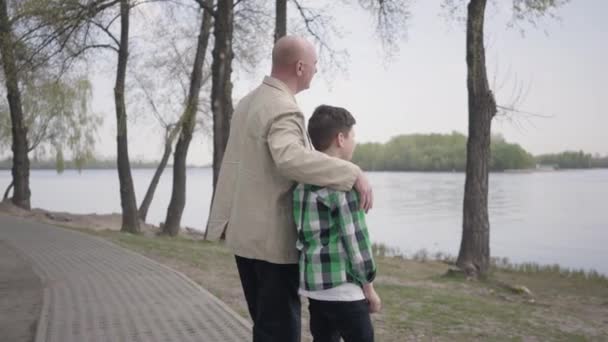 Grand-père et petit-fils debout sur la rive, regardant l'eau. L'homme serre l'enfant par les épaules. Concept des générations. Loisirs d'été. Connexion avec la nature — Video