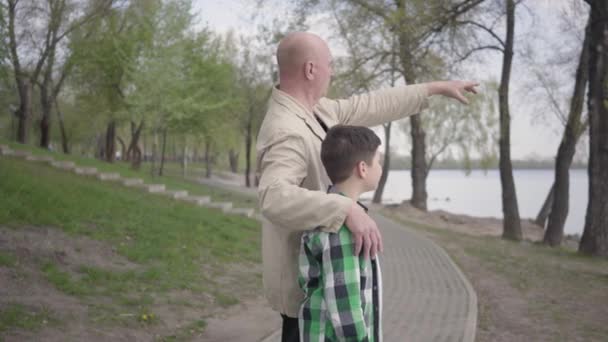 Abuelo y nieto de pie en la orilla del río, mirando el agua, apuntando a la distancia. El hombre abraza al niño por sus hombros. Concepto de generaciones. Tiempo libre de verano. Conexión con la naturaleza — Vídeos de Stock