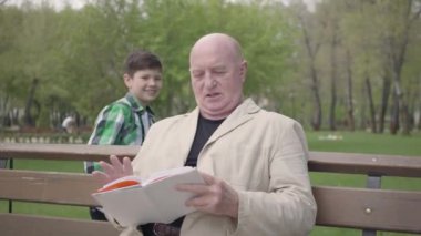 Portre sevimli kel olgun adam bankta parkta oturan bir kitap okuma. Arkadan gelen ve erkek omuzlarına el koyan sevimli küçük çocuk. Büyükbaba korkar, sonra torunukucaklar