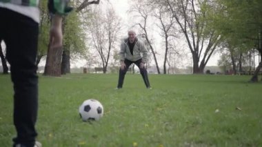 Torun topa vuruyor ve dedesi golü kaçırıyor. Mutlu sevimli çocuk futbol topu için yürürken ön planda atlama. Dışarıda eğlenen aile, aktif yaşam tarzı