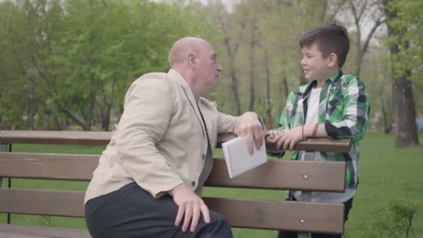 De kale volwassen man zittend in het Park op de Bank met een boek, praten met zijn kleinzoon. Dan schudden mensen de handen en lopen ze samen weg. Vrije tijd buitenshuis — Stockvideo