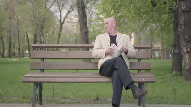The bald mature man sitting in the park on the bench reading a book. A little boy comes to him, the grandpa takes the hand of the kid and they walk away. Leisure outdoors, summertime fun — Stock Video