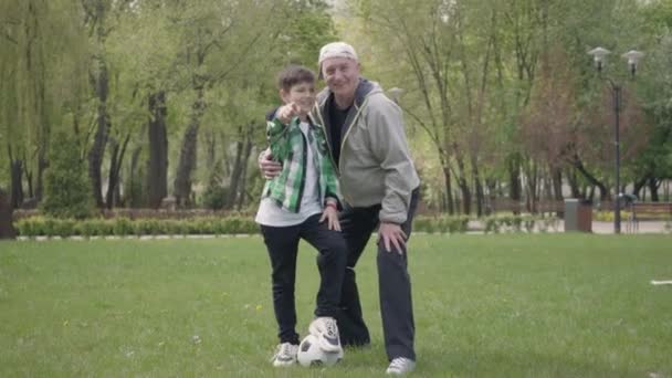 Retrato de felices y alegres abuelos y nieto de pie frente a la cámara y saludando con las manos. Nieto y abuelo después de jugar al fútbol abrazos mirando a la cámara al aire libre . — Vídeos de Stock