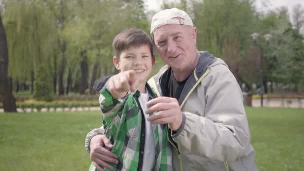 Portrait de mignon petit garçon heureux dans une chemise à carreaux et vieil homme positif regardant dans la caméra, invitant à les rejoindre dans le parc. La famille s'amuse dehors. Loisirs d'été — Video