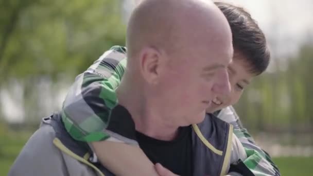 El hombre calvo maduro sentado en el parque en el banco leyendo el libro, niño pequeño abrazándolo por detrás. Ocio al aire libre. Abuelo y nieto pasando tiempo juntos — Vídeo de stock