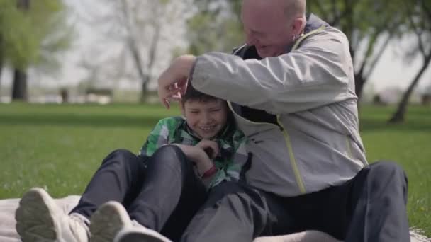 Retrato lindo hombre mayor sentado con su nieto en la manta en el parque, cosquillas al niño pequeño. El niño se ríe. Ocio al aire libre. Abuelo y nieto pasando tiempo juntos . — Vídeo de stock