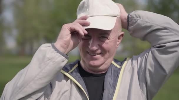 ポジティブな成熟した男は、公園で頭の上に帽子を置き、カメラを見て、笑顔。屋外で休んでいる老人。夏のレジャー — ストック動画