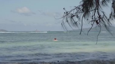 Seyşeller. Praslin Adası. Arka planda uzak larda berrak mavi suda sevimli sarışın kadın. Ön planda tropikal ağaç dalı. Güzel deniz manzarası. Tropikal ada lüks tatil.