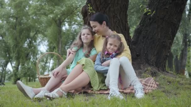 Retrato mulher sênior beijando e abraçando netas bonitos sentado na grama sob a árvore no parque, olhando em câmera sorrindo. Família feliz no piquenique. Lazer ao ar livre — Vídeo de Stock