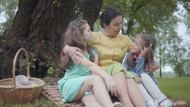 Ritratto elegante donna anziana seduta sull'erba sotto l'albero nel parco che abbraccia due graziose nipoti, raccontando loro una storia interessante. Famiglia felice al picnic. Tempo libero all'aperto — Video Stock