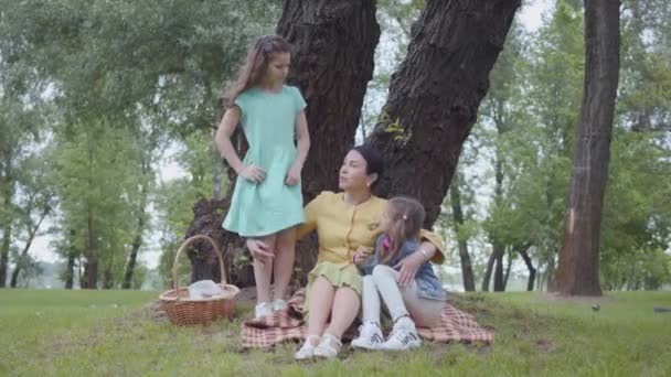 Елегантна старша жінка сидить на ковдрі під деревом у парку, розмовляючи з двома милими онуками. Одна дівчина сидить поруч, інша стоїть. Щаслива родина на пікніку. Дозвілля на відкритому повітрі — стокове відео