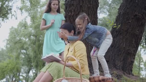 Mujer mayor elegante lindo sentado en la manta debajo del árbol en el parque leyendo el libro. Dos lindas nietas jugando cerca, una de ellas cubre los ojos de la mujer, tratando de llamar la atención — Vídeo de stock