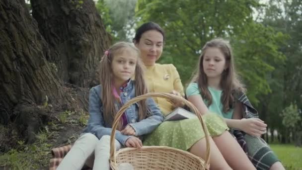 Mujer madura sentada en la hierba bajo el árbol en el parque con dos nietas lindas. Abuela sacando manzanas de la canasta y dándoselas a las niñas. Familia feliz pasar tiempo al aire libre — Vídeos de Stock