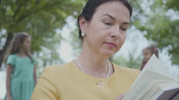 Portret van een elegante Senior grootmoeder die het boek in het Park op de voorgrond leest. Vervaagde figuren van twee die kleindochters op de achtergrond spelen. Gelukkige familie rust buitenshuis — Stockvideo