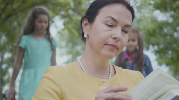 Portret van een elegante Senior vrouw die het boek in het Park op de voorgrond leest. Twee meisjes sluipen achter, één bedekt ogen van oma. Vrouw knuffelen kleindochters. Gelukkige familie rust buitenshuis — Stockvideo