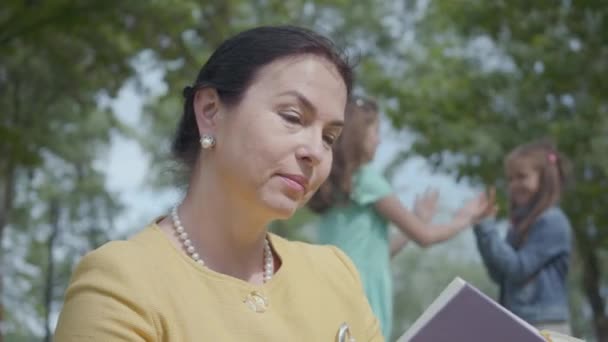 Retrato de una hermosa mujer madura linda leyendo un libro y sus adorables nietas juegan juntas en el parque al fondo. Feliz familia pasando tiempo al aire libre. Concepto de generaciones — Vídeos de Stock