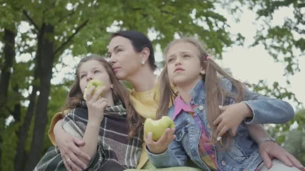 Mogen kvinna sitter under träden i parken med två söta barnbarn. Flickor som äter äpplen tittar bort. Lycklig familj spenderar tid utomhus. Hälsosam livsstil — Stockvideo
