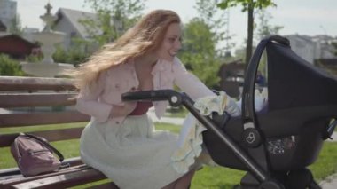 Genç mutlu kızıl saçlı anne, bebek arabasıyla evin yanındaki bankta oturuyor ve güzel bir bahar gününde gülümsüyor ve onunla konuşuyor.