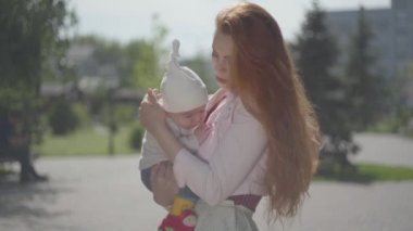Genç, güzel, kızıl saçlı anne kucağında sevimli bir bebek tutuyor ve güneşli bir bahar gününde onunla konuşuyor. Mutlu aile.