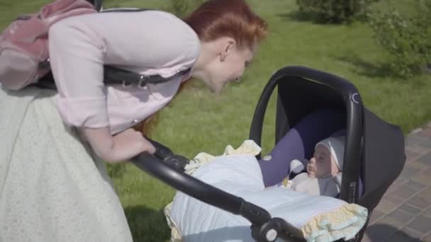 Kızıl saçlı anne parkta bir bebek arabasının üzerine eğilmiş. Bayan, dışarıda bebeğiyle güneşli bir günün tadını çıkarıyor. Genç anne bir çocukla yürüyor — Stok video