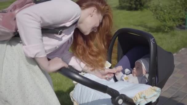 Όμορφη κοκκινομάλλα γυναίκα σκύψιμο ένα καροτσάκι στο πάρκο. Η κυρία απολαμβάνει την ηλιόλουστη μέρα με το μωρό της στην ύπαιθρο. Νεαρή μητέρα που περπατάει με ένα παιδί — Αρχείο Βίντεο