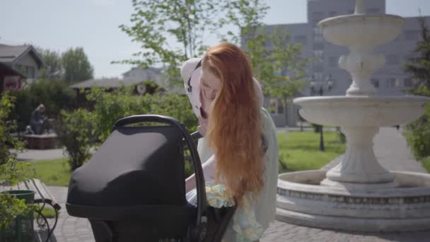 Hermosa mujer pelirroja inclinada sobre un cochecito y sonriendo en el parque. La señora disfrutando del día soleado con su bebé al aire libre. Madre joven caminando con un niño — Vídeos de Stock