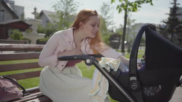 Schattig roodharige vrouw glimlachend kijken in de kinderwagen in het Park zittend op de Bank. De Vrouwe genieten van de zonnige dag met haar baby buitenshuis. Jonge moeder met een kind. Gelukkige familie — Stockvideo