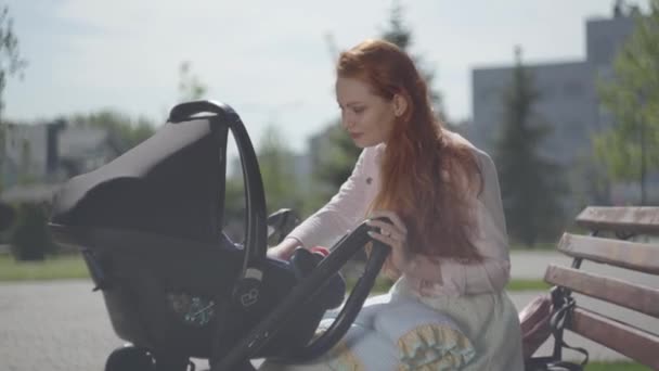 Roztomilá zrzavá žena, která si zahrává s dítětem, leží v kočárku v parku zblízka. Máma si užívala sluníčko se svým dítětem venku. Mladá matka s dítětem. Šťastná rodina — Stock video